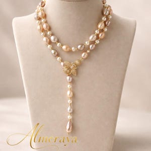 May include: A multi-strand pearl necklace with a gold-colored floral pendant and a single teardrop pearl. The necklace features pearls in shades of white, pink, and gold. The necklace is displayed on a mannequin.