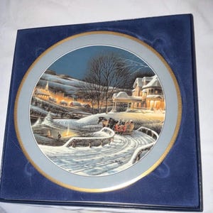 Puede incluir: Un plato decorativo con una escena invernal con un trineo tirado por caballos cruzando un puente nevado. El plato tiene un borde dorado y está colocado en una caja de terciopelo azul oscuro. La obra de arte representa un pueblo iluminado bajo un cielo nocturno.