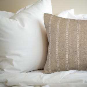 May include: A beige throw pillow with white stripes. The pillow is made of a textured fabric and has a subtle sheen.