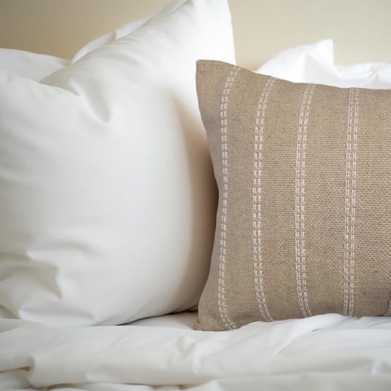 May include: A beige throw pillow with white stripes. The pillow is made of a textured fabric and has a subtle sheen.