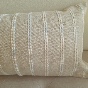 May include: A beige throw pillow with white stripes. The pillow is made of a textured fabric and has a simple, modern design.
