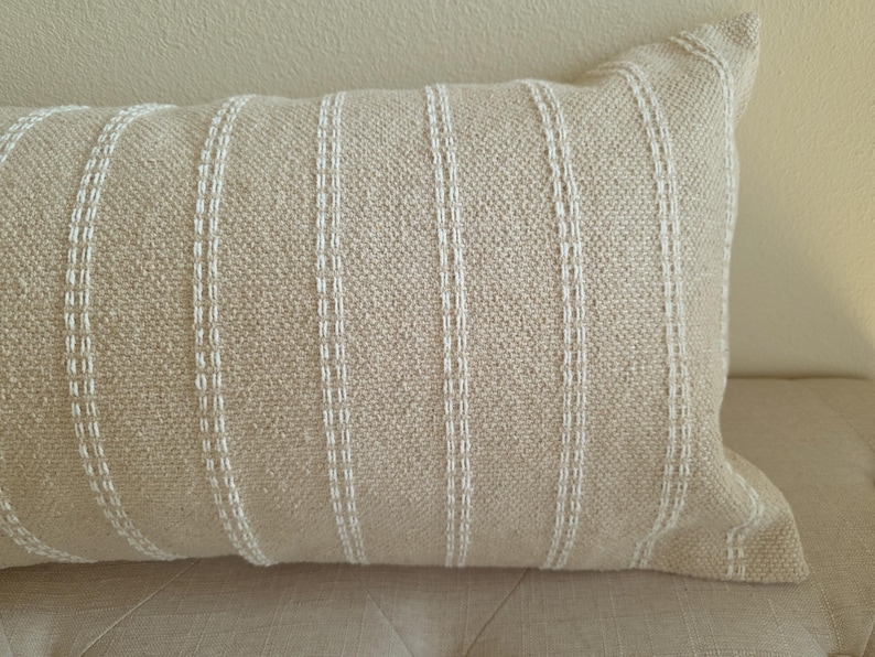 May include: A beige throw pillow with white stripes. The pillow is made of a textured fabric and has a simple, modern design.