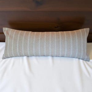 May include: A long, rectangular pillow with a light brown fabric and white vertical stripes. The pillow is lying on a white bedspread.
