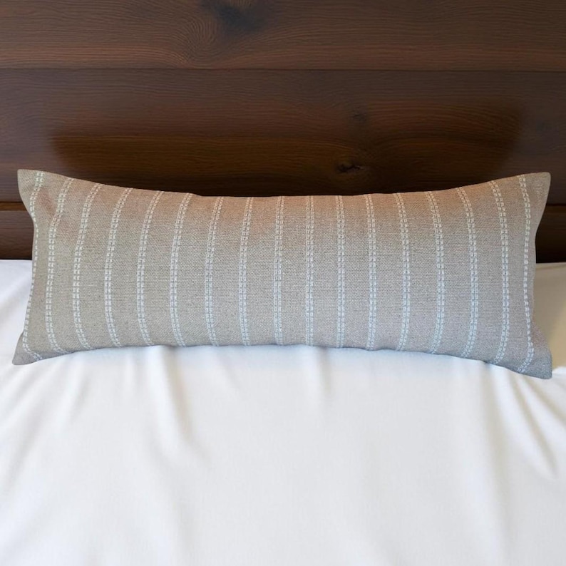 May include: A long, rectangular pillow with a light brown fabric and white vertical stripes. The pillow is lying on a white bedspread.