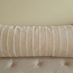 May include: A long, rectangular beige pillow with white stripes. The pillow is made of a textured fabric and has a subtle sheen.