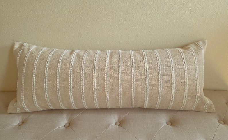 May include: A long, rectangular beige pillow with white stripes. The pillow is made of a textured fabric and has a subtle sheen.