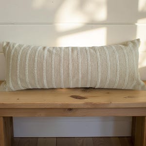 May include: A light beige lumbar pillow with white stripes sits on a wooden bench. The pillow is made of a soft, textured fabric.