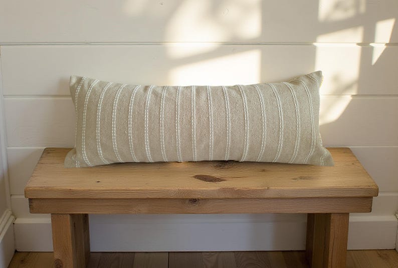May include: A light beige lumbar pillow with white stripes sits on a wooden bench. The pillow is made of a soft, textured fabric.