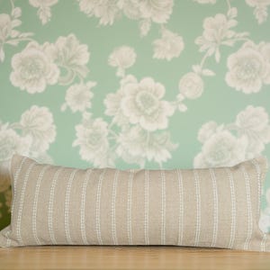May include: A long, rectangular, beige pillow with white stripes. The pillow is lying on a wooden surface in front of a green wall with a white floral pattern.