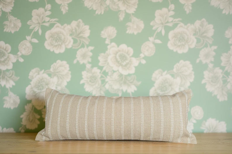 May include: A long, rectangular, beige pillow with white stripes. The pillow is lying on a wooden surface in front of a green wall with a white floral pattern.