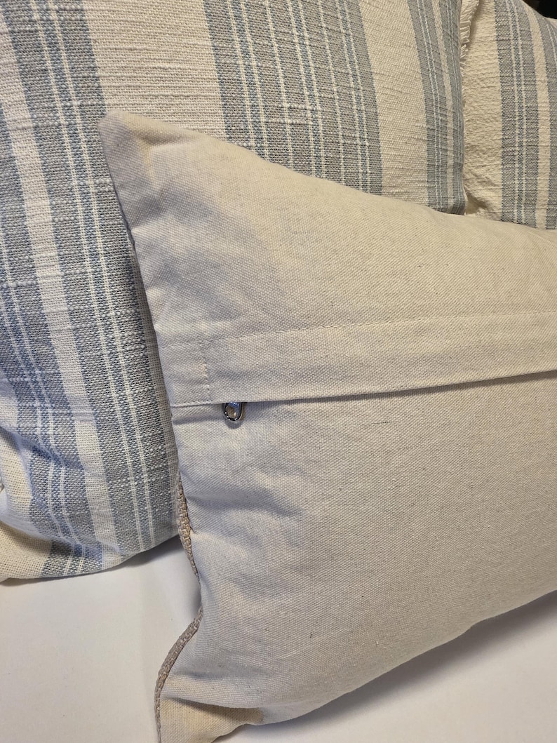 May include: Two throw pillows, one with a white and blue striped pattern and the other a solid white. The white pillow has a zipper closure on the back.