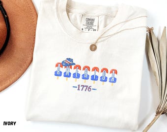 May include: Ivory-colored t-shirt with the word "AMERICA" in red and blue popsicle-style letters, a hat graphic, and the number "1776". The shirt has a crew neck and a small tag at the neckline.