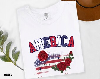 May include: White t-shirt with the word "AMERICA" in red and blue collegiate lettering. Below the word is a design of the American flag with red roses and the year "1776". The shirt is a Comfort Colors brand.