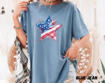 May include: A blue jean colored t-shirt featuring a star design with the American flag. The star is red, white, and blue with a watercolor effect. The text "BLUE JEAN" is at the bottom right of the image.
