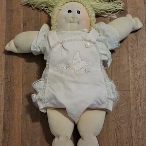May include: A soft fabric doll with pale skin, round cheeks, and blue eyes. It has light yellow yarn hair and wears a white short-sleeved shirt under white overalls featuring a butterfly embroidery on the chest. The doll wears white shoes.
