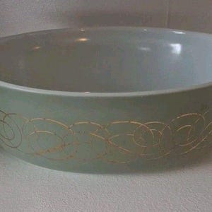May include: A round, light green glass casserole dish with integrated side handles. The exterior features a repeating gold-colored pattern of interconnected, swirling circular lines that wrap around the center of the bowl.
