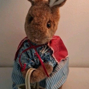 May include: A brown plush rabbit toy with long ears and dark eyes. The rabbit wears a blue floral blouse, a red cape with white lace trim, and a blue and white striped skirt. It holds a small, woven, light-colored basket with a handle.