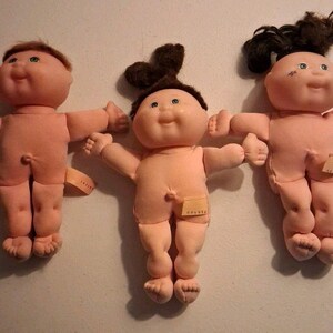 May include: Three soft-bodied dolls with vinyl heads and yarn hair. The left doll has short brown hair, the center doll has dark brown hair in a top knot, and the right doll has dark brown hair in a ponytail. Each doll has a small fabric tag on the hip.