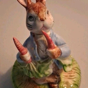 May include: A ceramic figurine of a brown rabbit wearing a light blue jacket. The rabbit is standing on a textured green base and holding two orange carrots in its paws. The rabbit has long ears, a white chest, and a detailed facial expression.