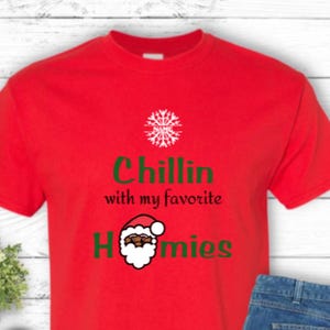 K&ouml;nnte beinhalten: Ein rotes T-Shirt mit dem Text "Chillin with my favorite Homies" und einer Weihnachtsmann-Grafik. Das Shirt wird mit einer blauen Jeans und schwarz-wei&szlig;en Turnschuhen kombiniert. Eine kleine gr&uuml;ne Pflanze ist ebenfalls im Bild.