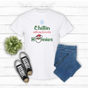 K&ouml;nnte beinhalten: Wei&szlig;es T-Shirt mit dem Text "Chillin with my favorite homies" in Gr&uuml;n und Rot. Das Design beinhaltet eine Schneeflocke und eine Weihnachtsmann-Grafik. Das Bild zeigt auch blaue Jeans und schwarze Leinenschuhe.