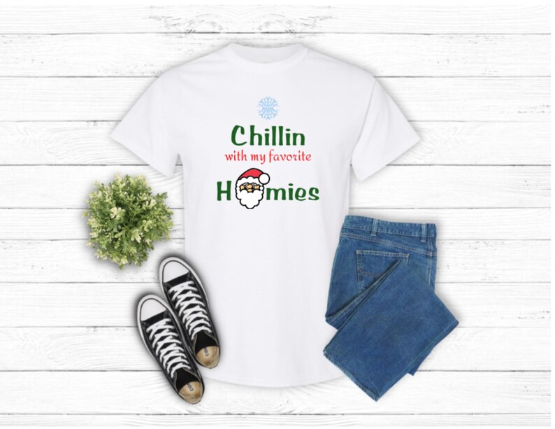 K&ouml;nnte beinhalten: Wei&szlig;es T-Shirt mit dem Text "Chillin with my favorite homies" in Gr&uuml;n und Rot. Das Design beinhaltet eine Schneeflocke und eine Weihnachtsmann-Grafik. Das Bild zeigt auch blaue Jeans und schwarze Leinenschuhe.