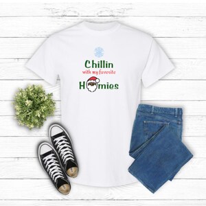 K&ouml;nnte beinhalten: Wei&szlig;es T-Shirt mit dem Text "Chillin with my favorite Homies" und einer Weihnachtsmann-Grafik. Das Shirt wird mit einer blauen Jeans und schwarz-wei&szlig;en Sneakers kombiniert. Eine kleine gr&uuml;ne Pflanze ist ebenfalls im Bild.