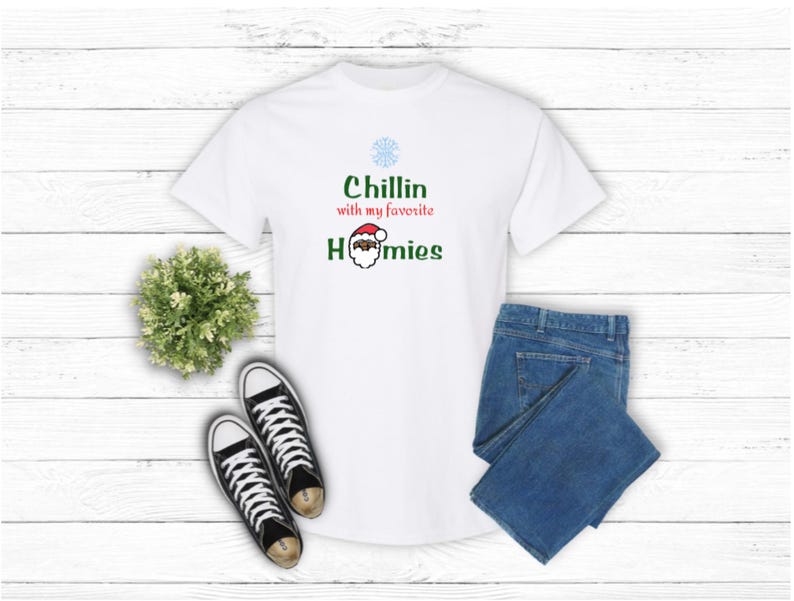 K&ouml;nnte beinhalten: Wei&szlig;es T-Shirt mit dem Text "Chillin with my favorite Homies" und einer Weihnachtsmann-Grafik. Das Shirt wird mit einer blauen Jeans und schwarz-wei&szlig;en Sneakers kombiniert. Eine kleine gr&uuml;ne Pflanze ist ebenfalls im Bild.