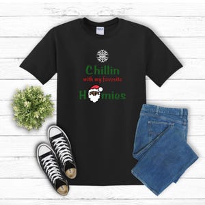 K&ouml;nnte beinhalten: Schwarzes T-Shirt mit dem Text "Chillin with my favorite Homies" in Gr&uuml;n und Rot. Das T-Shirt zeigt eine Weihnachtsmann-Grafik. Das Bild enth&auml;lt auch blaue Jeans und schwarz-wei&szlig;e Turnschuhe.