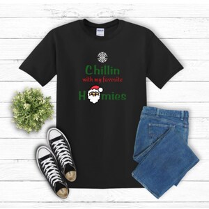 K&ouml;nnte beinhalten: Schwarzes T-Shirt mit dem Text "Chillin with my favorite Homies" in Gr&uuml;n und Rot, mit einer Weihnachtsmann-Grafik. Das Shirt wird mit blauen Jeans und schwarz-wei&szlig;en Turnschuhen kombiniert, vor einem wei&szlig;en Holzhintergrund.