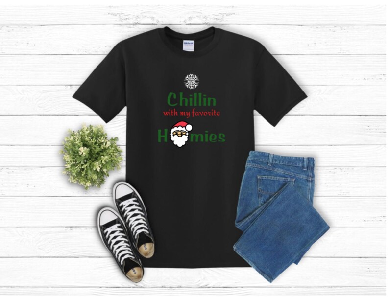 K&ouml;nnte beinhalten: Schwarzes T-Shirt mit dem Text "Chillin with my favorite Homies" in Gr&uuml;n und Rot, mit einer Weihnachtsmann-Grafik. Das Shirt wird mit blauen Jeans und schwarz-wei&szlig;en Turnschuhen kombiniert, vor einem wei&szlig;en Holzhintergrund.