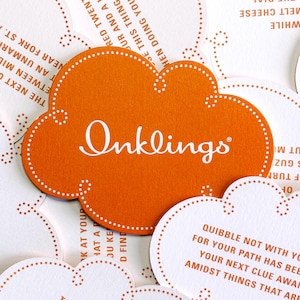 May include: A cloud-shaped orange card with the white text Inklings printed in the centre. The card features a white dotted border and is surrounded by other white cloud-shaped cards containing printed text in orange ink.