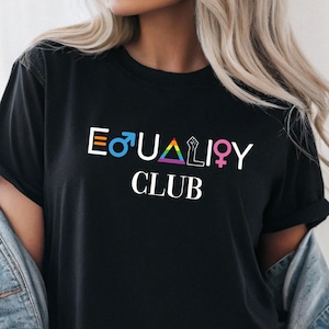 Könnte beinhalten: Schwarzes T-Shirt mit dem weißen und regenbogenfarbenen Schriftzug "EQUALITY CLUB". Das Design beinhaltet die männlichen und weiblichen Symbole, ein Regenbogen-Dreieck und eine geballte Faust.