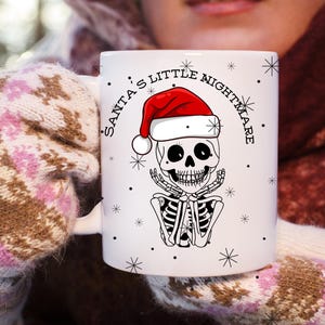 Könnte beinhalten: Weiße Keramik-Tasse mit einem Skelett, das einen Weihnachtsmannhut trägt. Die Tasse hat den Text "Santa's Little Nightmare" in einem geschwungenen Design, mit Schneeflocken und schwarzen Punkten. Das Skelett sitzt.