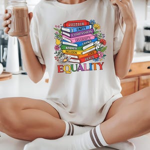 Könnte beinhalten: Cremefarbenes T-Shirt mit einem Stapel bunter Bücher mit Titeln wie "Gerechtigkeit" und "Gleichheit". Blumige Akzente umgeben die Bücher. Das Wort "EQUALITY" ist in Regenbogenfarben am unteren Rand des Designs gedruckt.