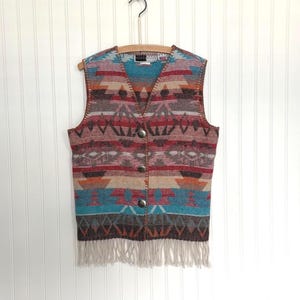 Puede incluir: Un chaleco estampado con cuello en V y flecos. El chaleco presenta un diseño geométrico en tonos azules, rojos, marrones y beige. Tiene tres botones plateados en la parte delantera y está colgado de una percha de madera.