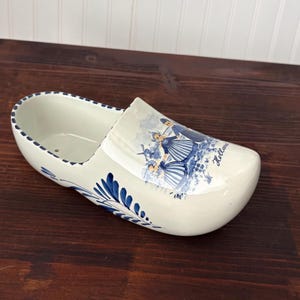 May include: A ceramic Dutch clog in white with blue floral and windmill designs. The clog has a rounded shape and a hollow interior. The word "Holland" is visible on the side. Decorative item.