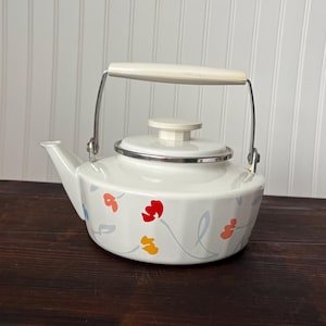 May include: White enamel teapot with a floral design in red, orange, and yellow. The teapot has a cream-colored handle and lid, and a silver rim. It sits on a dark wooden surface.