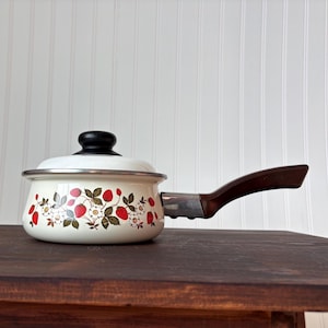 Op de afbeelding: Een vintage witte emaille steelpan met een deksel en een bruin handvat. De pot is versierd met een aardbeienpatroon in rood, groen en wit. De pot staat op een donkerbruin houten oppervlak.