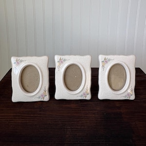 May include: Three off-white ceramic photo frames with oval apertures. Each frame has a square shape with rounded corners and a floral design in the upper corner. The frames are arranged on a dark brown wooden surface.