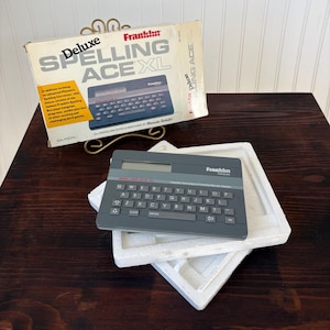 Puede incluir: Un corrector ortográfico electrónico Franklin Spelling Ace XL vintage. El dispositivo es gris con un teclado y una pequeña pantalla. La caja es beige y presenta el nombre y la descripción del producto.