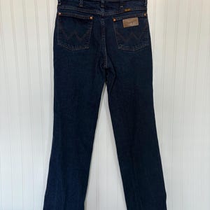 May include: Dark blue denim jeans with the Wrangler logo on the back pocket. The jeans are hanging on a metal hanger against a white background. The jeans have a classic five-pocket design.