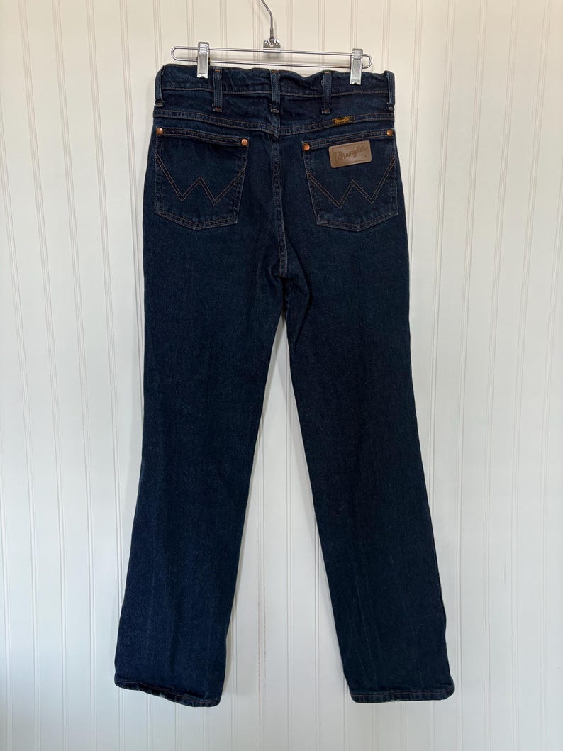 May include: Dark blue denim jeans with the Wrangler logo on the back pocket. The jeans are hanging on a metal hanger against a white background. The jeans have a classic five-pocket design.
