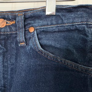 May include: Close-up of dark blue denim jeans. The jeans feature copper-colored rivets and a button, with visible stitching in a contrasting tan color. A small pocket is visible on the front.