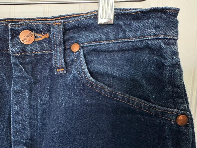 May include: Close-up of dark blue denim jeans. The jeans feature copper-colored rivets and a button, with visible stitching in a contrasting tan color. A small pocket is visible on the front.