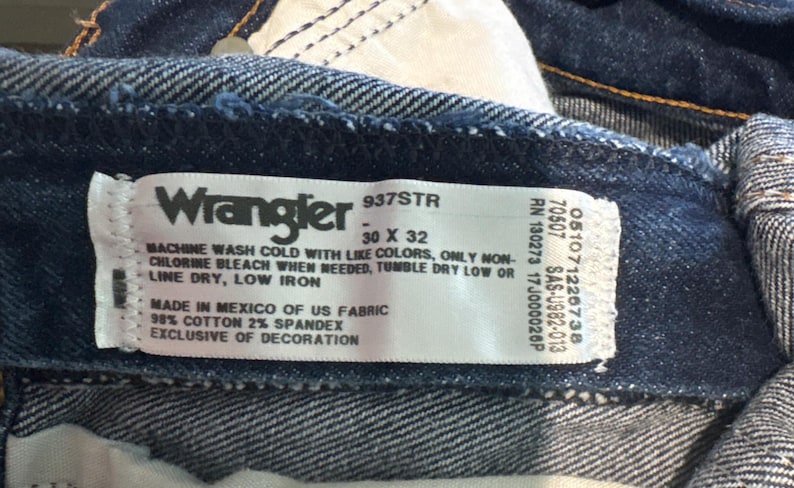 May include: Close-up of a Wrangler jeans label. The label is white with black text, indicating the brand, style, and size (30 x 32). It also includes washing instructions and fabric details: 98% cotton, 2% spandex.