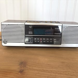 May include: A vintage GE clock radio in silver and black with a digital display. The radio has an AM/FM dial with frequency markings and a battery backup feature. The clock radio is sitting on a wooden surface.