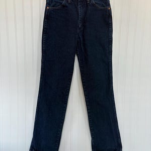 May include: Dark wash denim jeans hanging on a metal hanger. The jeans have a classic five-pocket design with copper rivets and a button closure. The straight-leg style is visible, and the jeans appear to be in good condition.