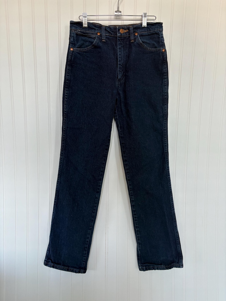 May include: Dark wash denim jeans hanging on a metal hanger. The jeans have a classic five-pocket design with copper rivets and a button closure. The straight-leg style is visible, and the jeans appear to be in good condition.