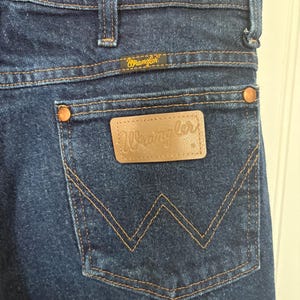May include: Close-up of dark blue denim jeans with the Wrangler logo on the back pocket and waistband. The jeans have copper-colored rivets and a classic five-pocket design. The stitching is a contrasting tan color.